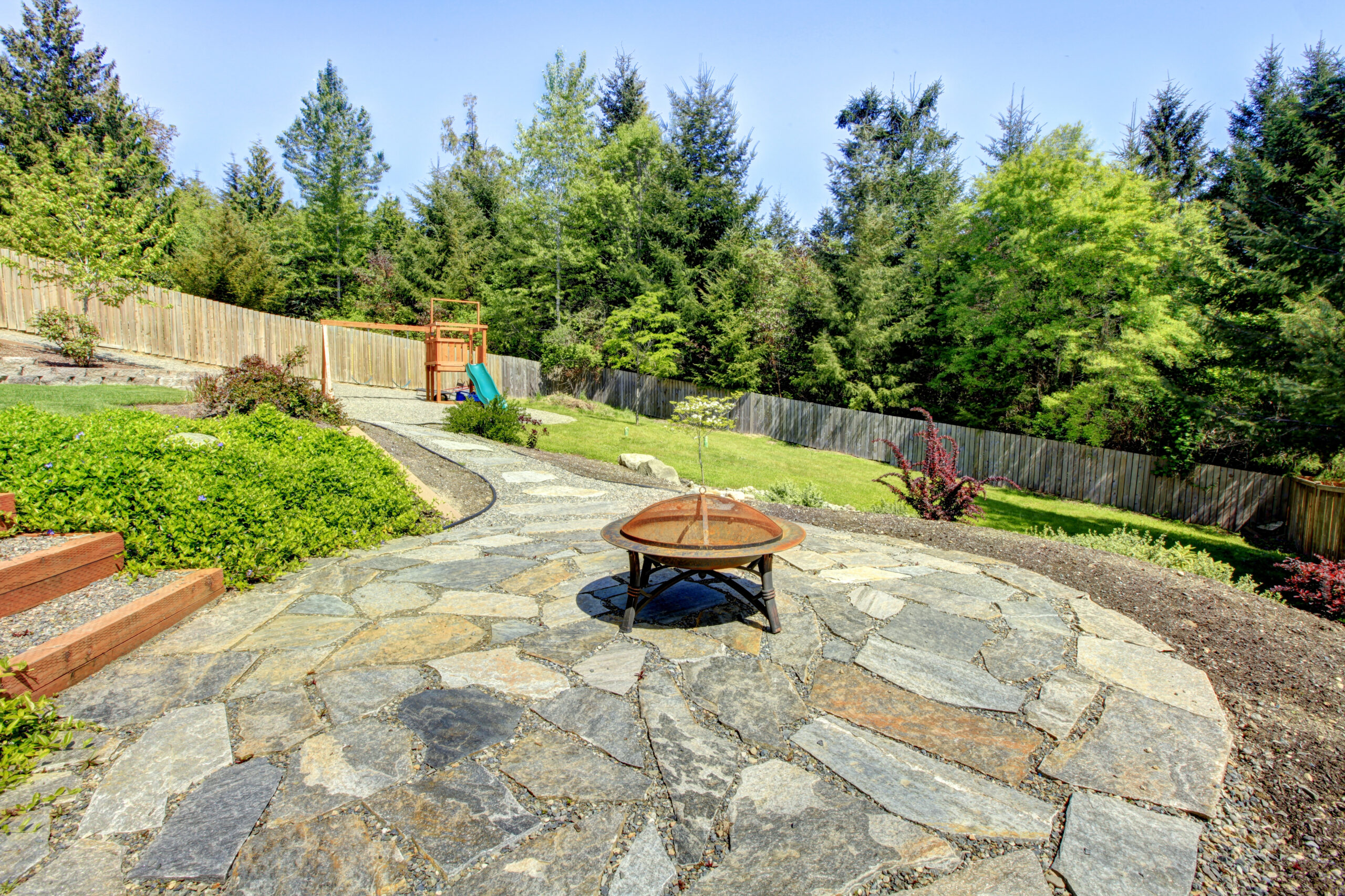 Large fenced backyard with stone and fire pit with trees.