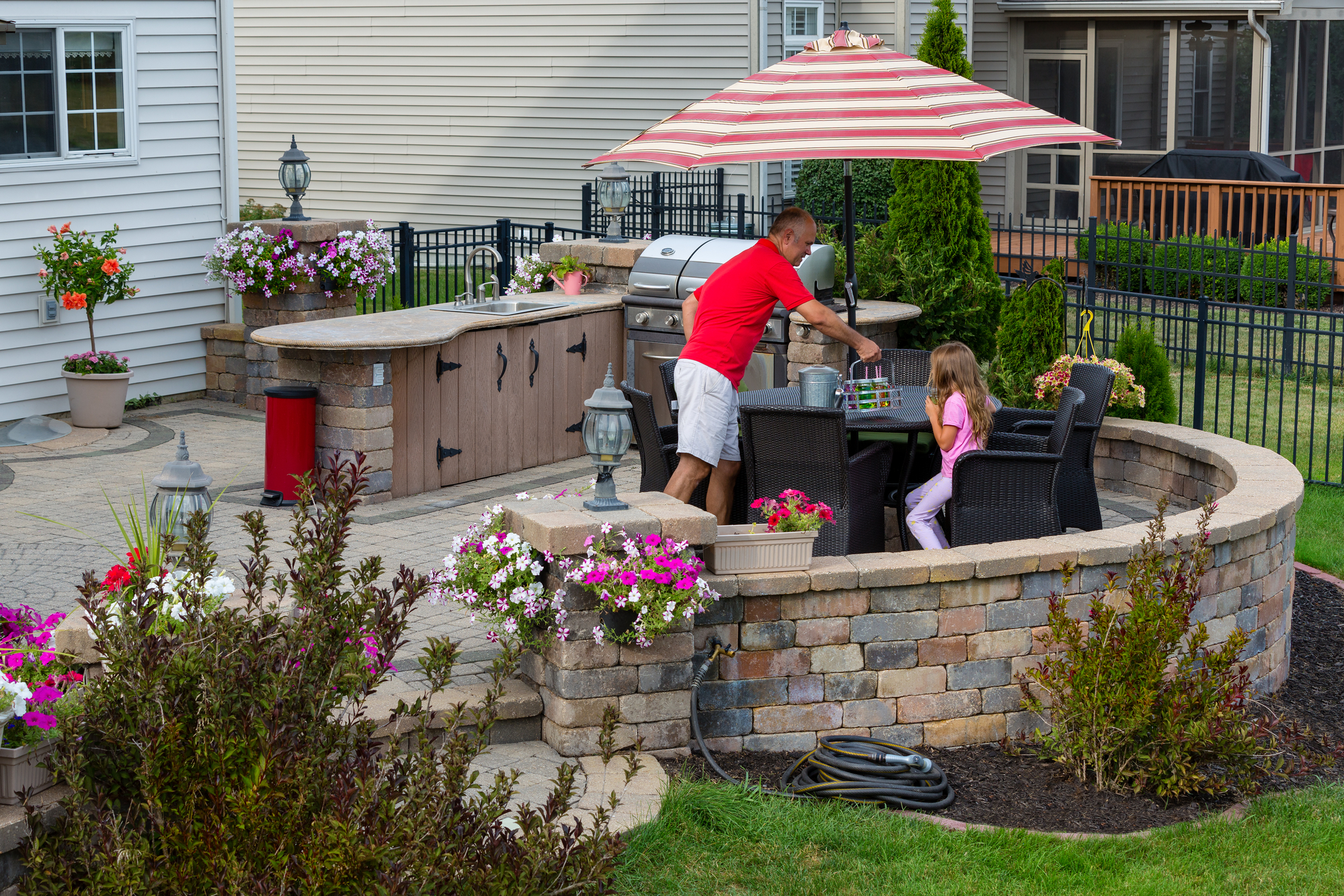 Daddy serving lemonade to his little daughter outdoors on a brick patio as she relaxes in the shade of an umbrella near an outdoor kitchen and gas barbecue in summer